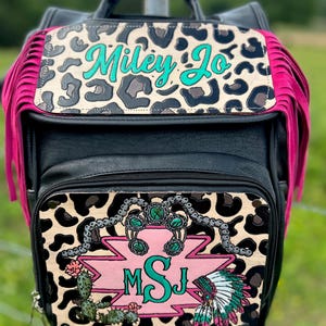 May include: Black backpack with a leopard print flap and pink fringe. The flap has the name "Miley Jo" in teal script. The front pocket features a pink and turquoise design with the initials "MSJ", a cactus, and a Native American headdress.