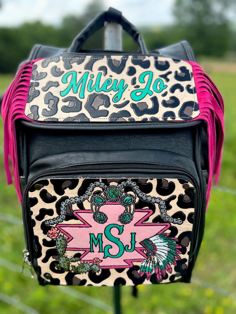 May include: Black backpack with a leopard print flap and pink fringe. The flap has the name "Miley Jo" in teal script. The front pocket features a pink and turquoise design with the initials "MSJ", a cactus, and a Native American headdress.