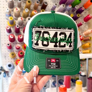May include: A green and white trucker hat featuring a rectangular patch with frayed edges. The patch displays the black text 76424 with the green cursive word Breckenridge embroidered over it. A black sticker on the brim reads 112 The Original MD-LG.