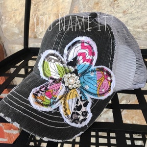 May include: A black and gray trucker hat with a large, colorful patchwork flower applique. The flower has a rhinestone center and is sewn onto the front of the hat.