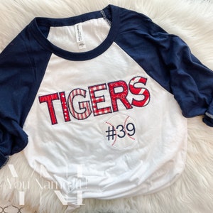 May include: White baseball tee with a red, white, and blue patchwork "TIGERS" applique and a baseball with the number "#39" on it. The shirt has a navy blue raglan sleeve.