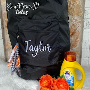 May include: Black drawstring bag with a zippered pocket and a white embroidered name "Taylor". The bag has a black and white checkered ribbon attached to it. A yellow bottle of Tide laundry detergent is next to the bag.