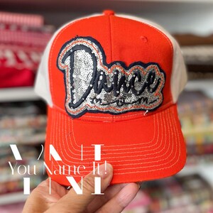 May include: A red and white trucker hat with the word "Dance" in silver sequins. The hat has a curved brim and a mesh back. The text "You Name It!" is visible at the bottom of the image.