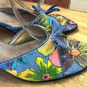 May include: A pair of floral-patterned, open-toe heels with a blue background and a small heel. The shoes have a bow detail on the front and clear straps. The floral pattern includes yellow, pink, and green flowers.