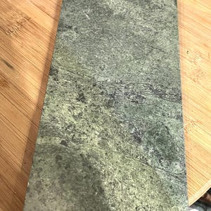 May include: A rectangular green marble serving board with a decorative silver-colored handle featuring an apple design. The board has a natural, mottled pattern and is ideal for serving appetizers or cheeses.