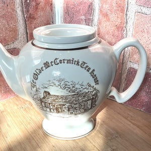 May include: A white ceramic teapot featuring a brown graphic illustration of a building and the text "Ye Olde McCormick Tea House" printed on the front. The teapot has a curved handle, a pointed spout, a circular base, and a removable lid on top.