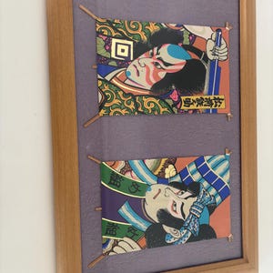 May include: Framed artwork featuring two Japanese Ukiyo-e prints. Each print depicts a stylized Kabuki actor with vibrant colors, intricate patterns, and Japanese text. The prints are mounted on a purple background within a wooden frame.