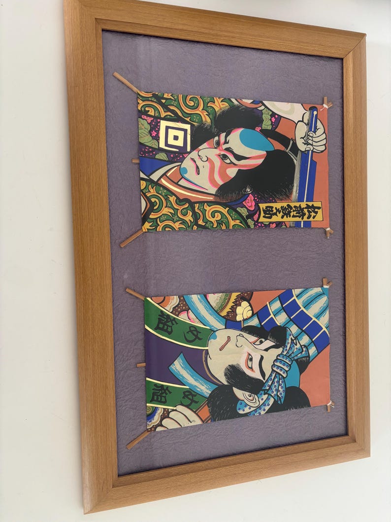 May include: Framed artwork featuring two Japanese Ukiyo-e prints. Each print depicts a stylized Kabuki actor with vibrant colors, intricate patterns, and Japanese text. The prints are mounted on a purple background within a wooden frame.