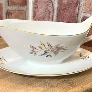 May include: White porcelain gravy boat and saucer set. The gravy boat has a curved handle and a floral design with gold trim. The saucer also has a gold trim and a matching floral design. The set is on a wooden surface.