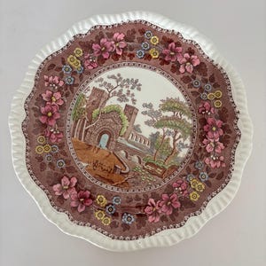 May include: A round, vintage decorative plate with a scalloped edge. The plate features a central landscape scene surrounded by a brown border with pink, yellow, and blue floral designs. The rim is white.