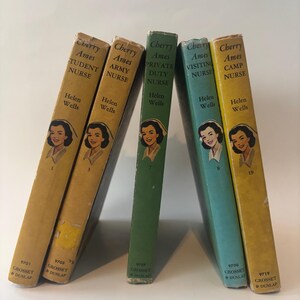 May include: A collection of vintage "Cherry Ames" novels by Helen Wells. The books, in various colors, feature a portrait of a nurse on the cover. Titles include "Student Nurse," "Army Nurse," and "Camp Nurse."