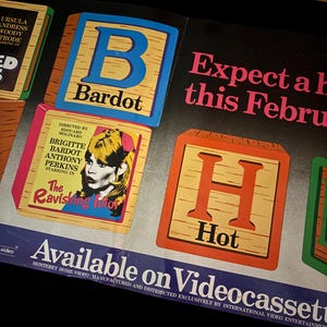 May include: Vintage movie poster featuring Brigitte Bardot. The poster includes the text "The Ravishing Idiot" and "Hot" on wooden blocks. The poster also states "Available on Videocassette."
