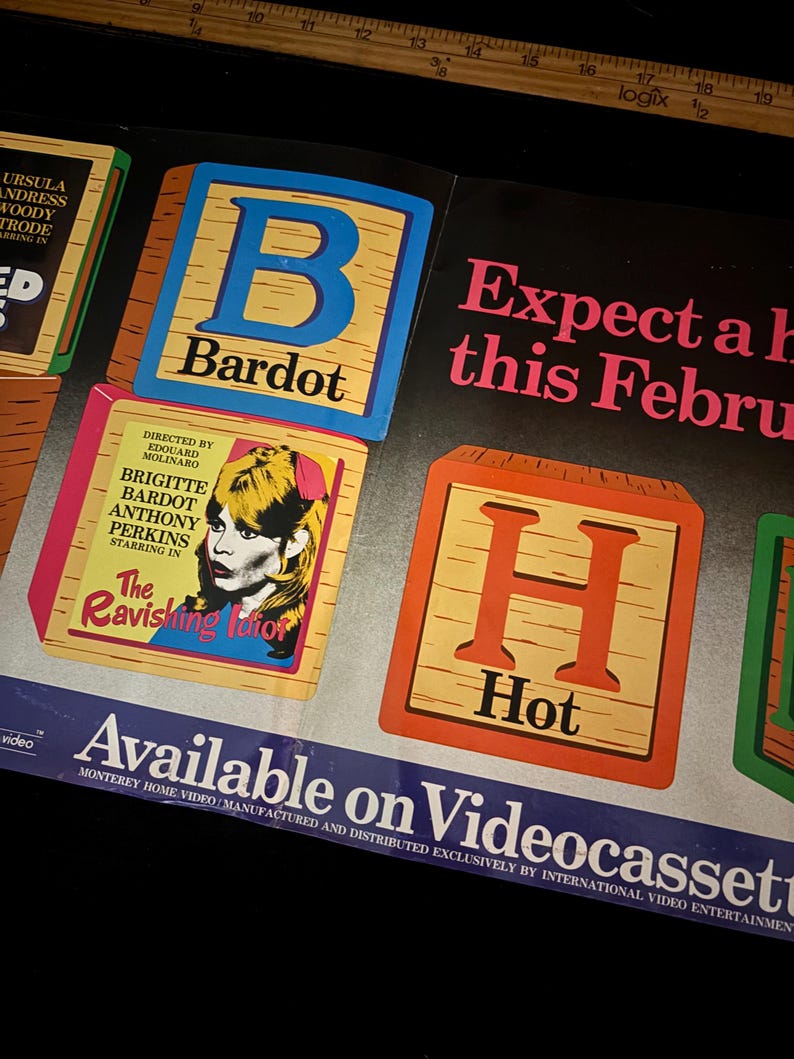May include: Vintage movie poster featuring Brigitte Bardot. The poster includes the text "The Ravishing Idiot" and "Hot" on wooden blocks. The poster also states "Available on Videocassette."