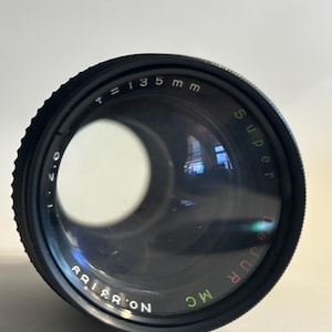 May include: A black camera lens with the text "T=135mm" and "Super Dejur MC". The lens has a large, clear glass element and a ridged focusing ring. The lens has the number "NO. 83189" printed on it.