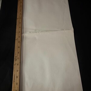 May include: A folded, off-white sheet of paper with a wooden yardstick along the left edge. The yardstick is marked with inch measurements. The paper is on a black surface.
