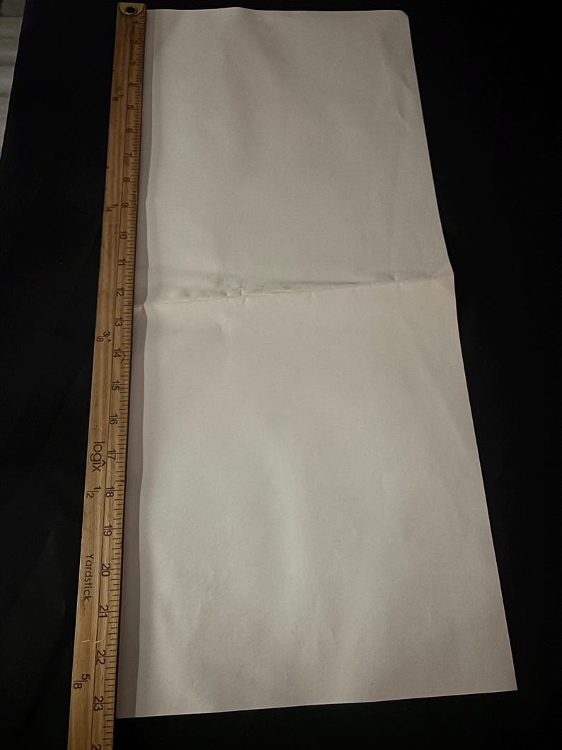 May include: A folded, off-white sheet of paper with a wooden yardstick along the left edge. The yardstick is marked with inch measurements. The paper is on a black surface.