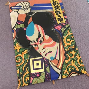 May include: A framed Japanese woodblock print featuring a Kabuki actor. The print showcases a detailed illustration with vibrant colors, including red, blue, and green. The artwork is mounted on a purple background and held in place with small wooden dowels.