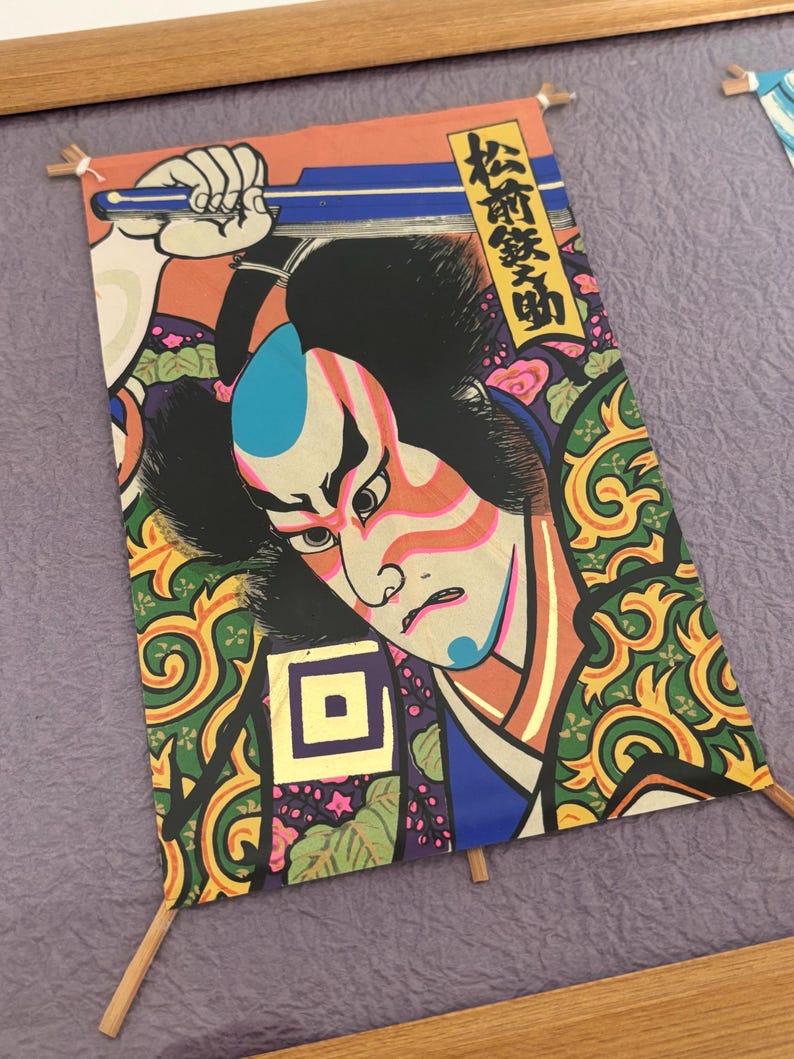 May include: A framed Japanese woodblock print featuring a Kabuki actor. The print showcases a detailed illustration with vibrant colors, including red, blue, and green. The artwork is mounted on a purple background and held in place with small wooden dowels.