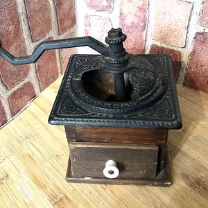 May include: Antique manual coffee grinder with a dark metal top and handle, and a wooden base with a drawer. The grinder has an ornate design on the top and a wooden handle. The drawer has a white knob.