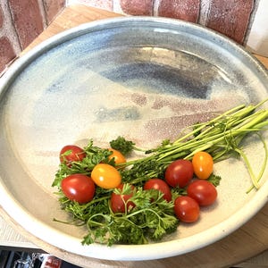 May include: A large, round ceramic serving platter with a light blue and gray painted design. The platter contains red and yellow cherry tomatoes, green parsley, and green stalks. The platter is on a wooden surface.