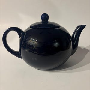 May include: A glossy, dark navy blue ceramic teapot with a rounded body, a curved handle, a matching lid with a small knob handle, and a short, tapered spout. The surface of the teapot is smooth and reflective, showing light highlights.