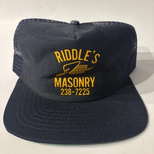 May include: A navy blue trucker hat with a mesh back and a flat brim. The front panel features yellow screen-printed text that reads RIDDLE'S MASONRY 238-7225, accompanied by a graphic illustration of a masonry trowel and a stack of bricks.