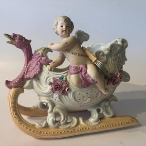 May include: A porcelain figurine of a cherub sitting in a decorative sleigh. The sleigh features a pink bird-like figure at the front, ornate scrollwork, and applied floral details. The cherub wears a pink sash and carries a quiver of arrows.