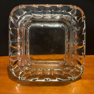 Op de afbeelding: Een vierkante asbak van helder glas met een verhoogde, getextureerde rand. De asbak heeft een glad, vlak interieur.