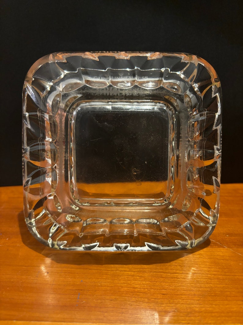 Op de afbeelding: Een vierkante asbak van helder glas met een verhoogde, getextureerde rand. De asbak heeft een glad, vlak interieur.