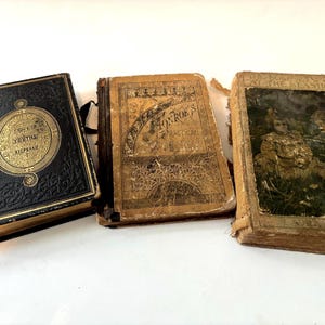 May include: Three antique books with worn covers. The first book is black with gold lettering and a decorative border. The second book is tan with text and a decorative border. The third book has a color illustration on the cover.