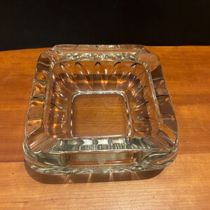 Op de afbeelding: Een vierkante asbak van helder glas met een decoratief geribbeld ontwerp rond de randen.