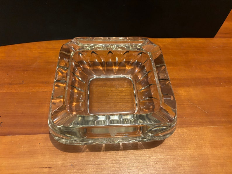 Op de afbeelding: Een vierkante asbak van helder glas met een decoratief geribbeld ontwerp rond de randen.