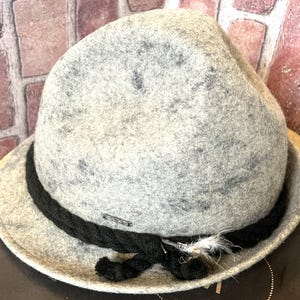 May include: A gray wool hat with a black braided band and a small feather accent. The hat has a speckled pattern and a classic fedora shape. A small metal bar is attached to the black band. This hat is a stylish accessory.