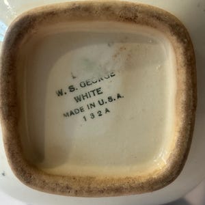 May include: The underside of a white ceramic vessel featuring a square base with a brown, unglazed rim. The center is stamped with black text reading W. S. GEORGE, WHITE, MADE IN U.S.A., and 132 A. The ceramic surface shows signs of age and wear.
