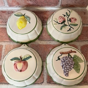 May include: Four white ceramic molds with green trim, each featuring a different fruit design: a lemon, raspberries, grapes, and an apple. The molds are perfect for making individual desserts.