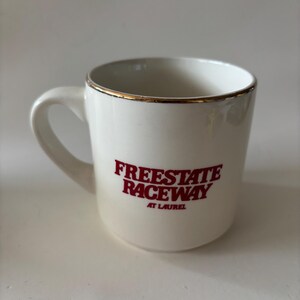 May include: White ceramic mug with a gold rim and a curved handle. The mug features the red text "FREESTATE RACEWAY AT LAUREL". The mug is designed for beverages and is suitable for home or office use.
