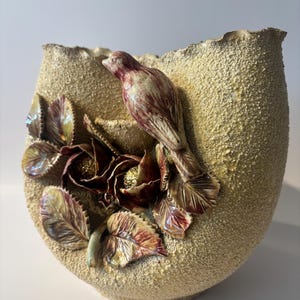 May include: A beige ceramic vase with a rough, textured surface. The front features a three-dimensional sculpted scene of a small bird perched above two dark red flowers with gold centers, surrounded by several detailed green and red leaves.