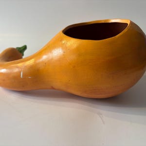 May include: A hollow, gourd-shaped ceramic vessel with a smooth, orange-colored exterior and a small, green-painted stem at the narrow end. The interior of the bowl is dark brown, and the overall form mimics the shape of a butternut squash.