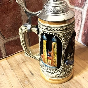 May include: A decorative beer stein with a silver lid and handle. The ceramic body features a detailed relief of a European cathedral with blue and yellow accents. The stein is set on a wooden surface.