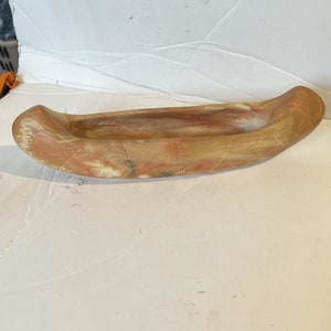 May include: A small, hand-crafted canoe made of clay. The canoe is painted in shades of brown, orange, and white.