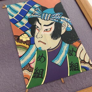 May include: Framed Japanese woodblock print featuring a Kabuki actor. The artwork displays vibrant colors, including orange, blue, green, and purple. The actor's face is detailed with traditional makeup, and Japanese characters are visible on the garment.