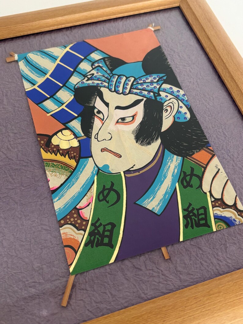 May include: Framed Japanese woodblock print featuring a Kabuki actor. The artwork displays vibrant colors, including orange, blue, green, and purple. The actor's face is detailed with traditional makeup, and Japanese characters are visible on the garment.