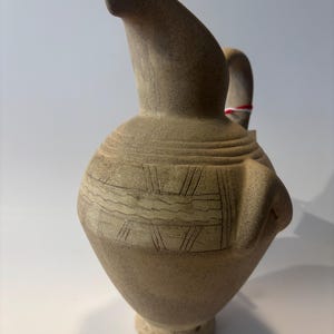 May include: A tan ceramic pitcher with a textured, matte finish. The vessel features a rounded body, a narrow neck with a flared spout, and a handle. The surface is decorated with horizontal bands and incised geometric patterns around the midsection.