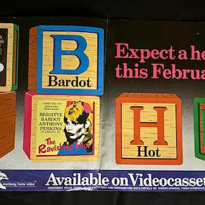 May include: Vintage movie advertisement featuring wooden blocks spelling out "ABH" and "M". The blocks display movie titles and actors, including "Loaded Guns" and "The Ravishing". The text "Expect a heat wave this February" is also visible.