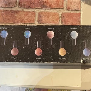 May include: A black box featuring illustrations of the planets in our solar system. Each planet is represented by a colorful sphere on a black stand. The box includes the names of the planets: Mercury, Earth, Venus, Mars, Jupiter, Saturn, Uranus, Neptune, and Pluto.