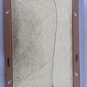 May include: Back of a framed artwork with a gold textured background. The frame is made of light brown wood. A black wire is strung across the back for hanging. White plastic clips and metal hardware secure the artwork.