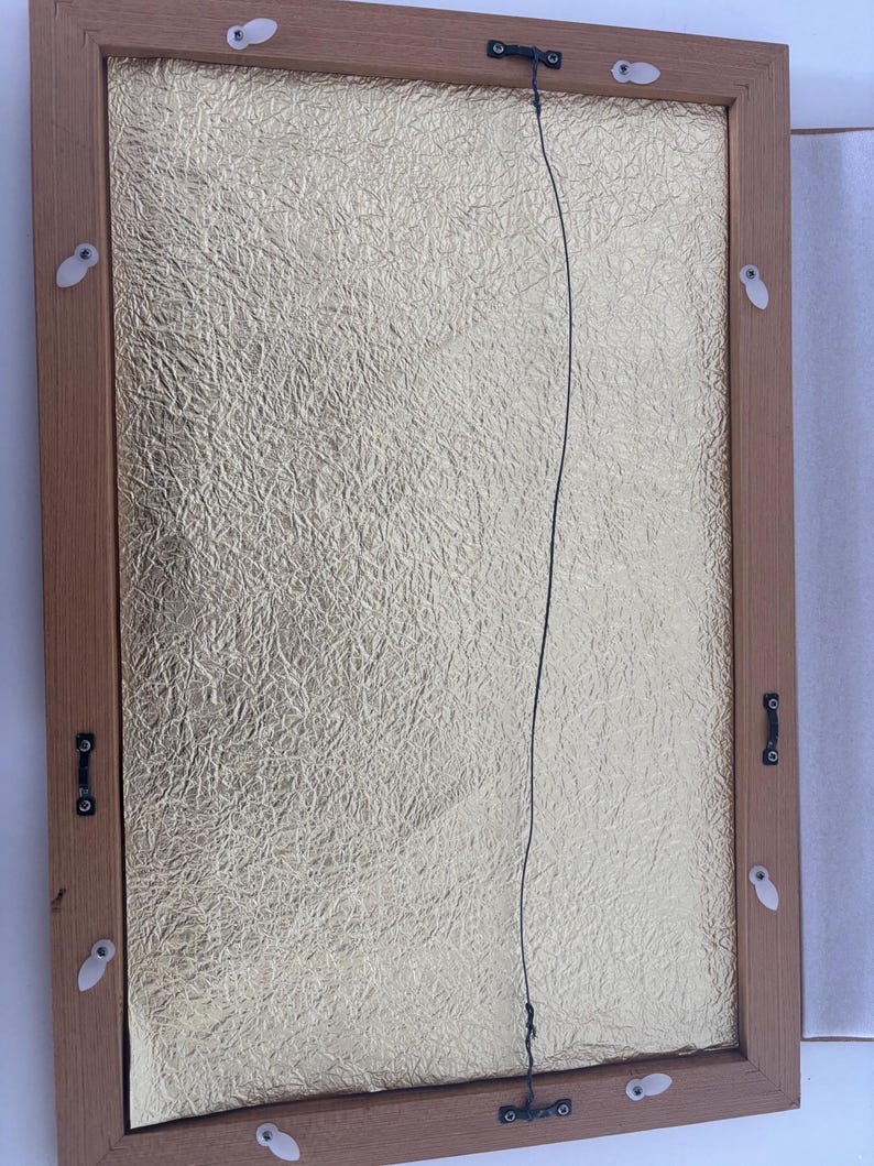 May include: Back of a framed artwork with a gold textured background. The frame is made of light brown wood. A black wire is strung across the back for hanging. White plastic clips and metal hardware secure the artwork.