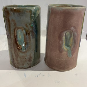 May include: Two ceramic mugs with a unique glaze pattern. The mugs are a light blue-green and a light pink-brown color. The glaze has a mottled effect with swirls of color.