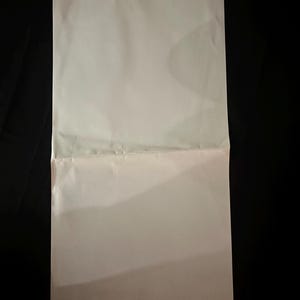 May include: A folded, off-white sheet of paper against a black background. The paper is creased horizontally, dividing it into two sections. The lighting creates shadows and highlights on the paper's surface.