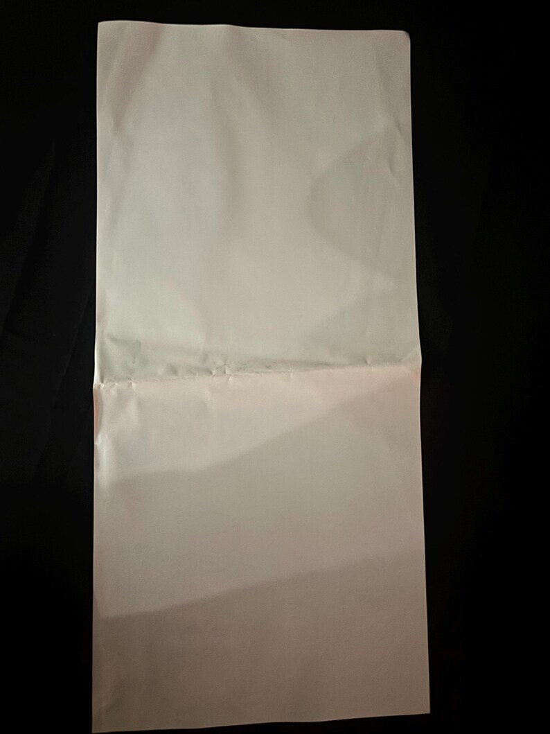 May include: A folded, off-white sheet of paper against a black background. The paper is creased horizontally, dividing it into two sections. The lighting creates shadows and highlights on the paper's surface.